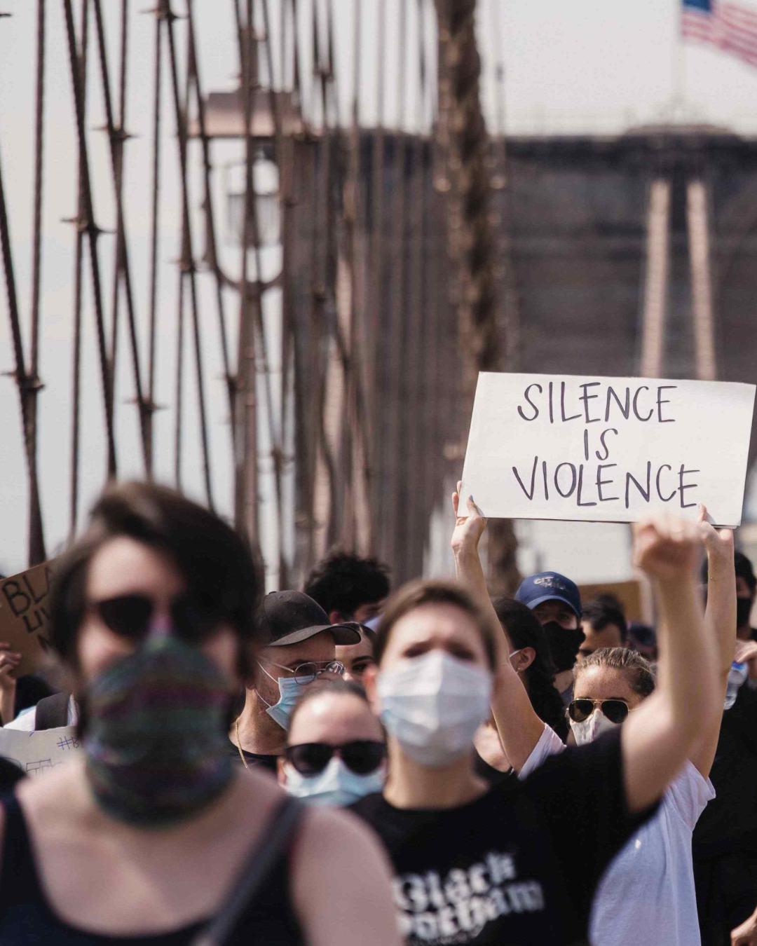 A crowd of individuals marching across a bridge. Some individuals hold signs that say "silence is violence" and "no justice no peace".